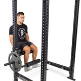 A man in a black shirt, black shorts, and white shoes prepares for a leg workout using the Bells of Steel Leg Curl / Leg Extension Rack Attachment on a power rack with weight plates.