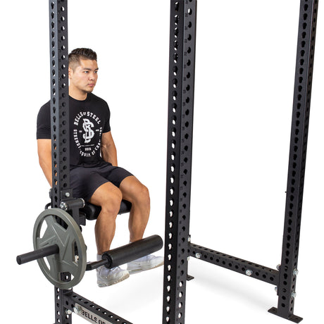 A man in a black T-shirt and shorts is seated on the Leg Curl / Leg Extension Rack Attachment by Bells of Steel, demonstrating an ideal home gym setup. The power rack, featuring various attachments and weights, stands out against the white background.