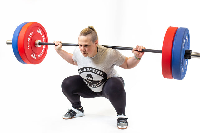 A person executes a barbell squat, lifting the "Multi-Purpose Olympic Barbell – The Utility Bar" and wearing a Bells of Steel shirt, black leggings, and lifting shoes against a white background.