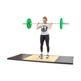 A woman with a ponytail performs a barbell squat on a lifting platform, wearing a logo T-shirt, black leggings, and weightlifting shoes. The barbell is loaded with Bells of Steel's Coloured Bumper Plates featuring stainless steel insert rings for precision and accurate weight distribution.