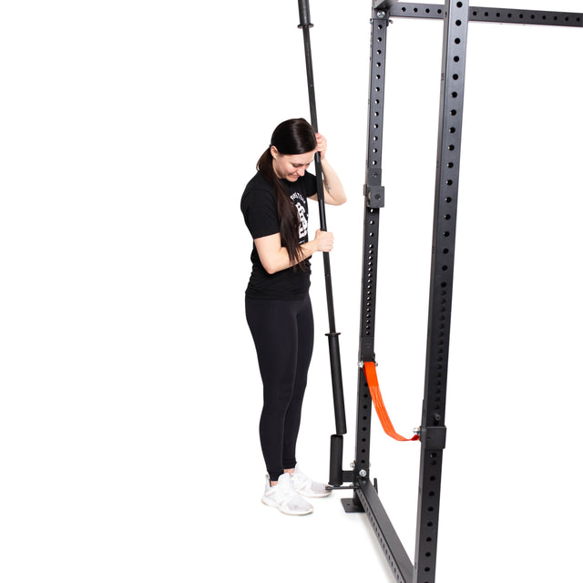 A woman dressed in black workout attire is adjusting a barbell on the Bells of Steel Vertical Mount Barbell Holder Rack Attachment. Set against a white background that highlights both the equipment and her actions, bright orange accents add a vibrant flair to the organization of the home gym.