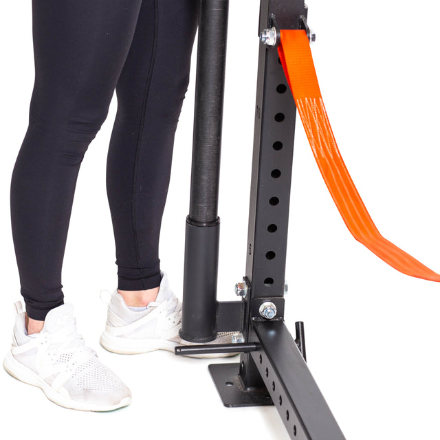 A person in black leggings and white sneakers stands next to a Bells of Steel Vertical Mount Barbell Holder Rack Attachment with an attached orange band. They're holding onto a cylindrical black grip on the frame. This setup is ideal for organizing your home gym against the plain white background.