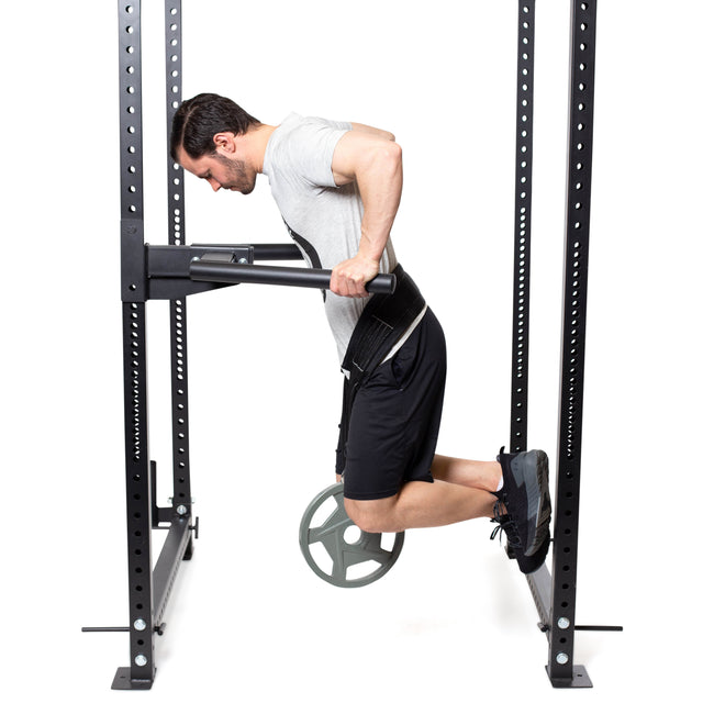 A man showcases great weight distribution doing weighted dips on a power rack, wearing a gray T-shirt and black shorts. He's using the Bells of Steel Belt Squat Belt to hold a plate, set against a plain white backdrop.