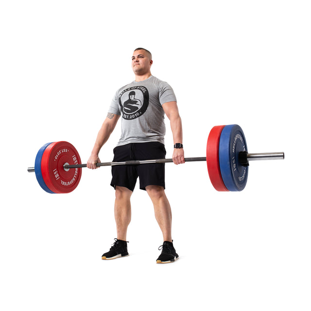 A person in a gray T-shirt and black shorts lifts a Bells of Steel barbell with Coloured Bumper Plates. The precise weight is visible in their stance, feet shoulder-width apart, looking forward. The shiny stainless steel insert contrasts with the white background.