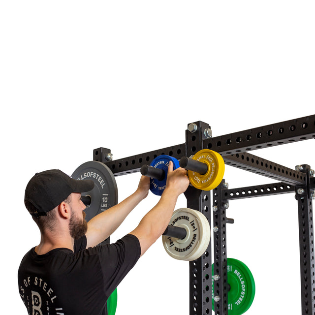 A person in a black cap and T-shirt adjusts Bells of Steel Change Plate Storage Pegs on a power rack with colorful weights and a pull-up bar, preparing for a workout against the plain white background.