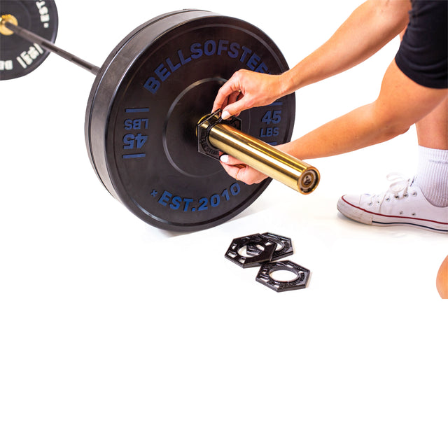 A person wearing white sneakers adjusts a weight on an Olympic barbell, which has cast iron plates marked "BELLS OF STEEL" and "45 LBS." Nearby are hexagonal collars.