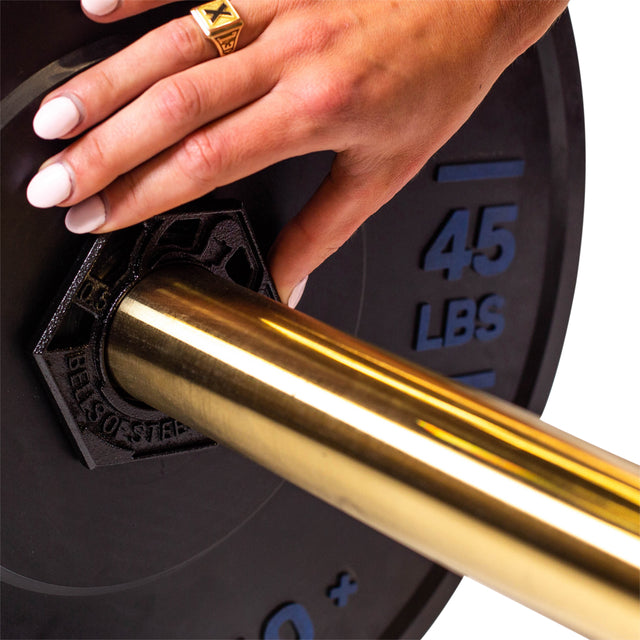 A hand with manicured nails and a gold ring is adjusting the Bells of Steel Fractional Iron Plate - 0.5 LB on an Olympic barbell with a golden finish.