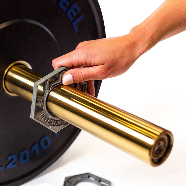 A hand secures a hexagonal weight collar labeled "Bells of Steel" onto a shiny, gold Olympic barbell with black plates. The background is white.