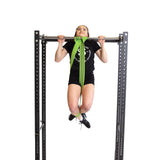 Wearing athletic gear, a person does a pull-up on a metal bar with a Bells of Steel Fabric Non-Slip Resistance Band (41")—green, looped over the bar and under their feet for support—against a plain white background.