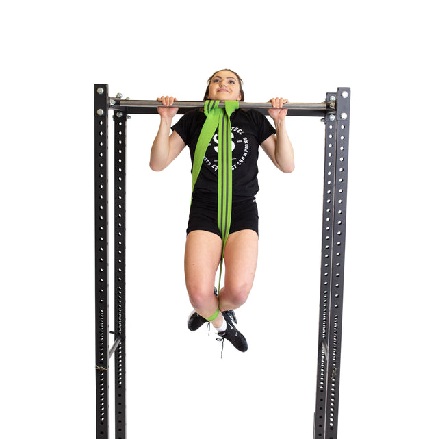 Wearing athletic gear, a person does a pull-up on a metal bar with a Bells of Steel Fabric Non-Slip Resistance Band (41")—green, looped over the bar and under their feet for support—against a plain white background.