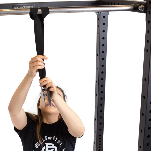 A person attaches a Bells of Steel Cable Pulley to a pull-up bar on a black metal power rack, showcasing its sturdy design. The individual, with long hair and a black shirt featuring a white logo, stands against a plain white background.