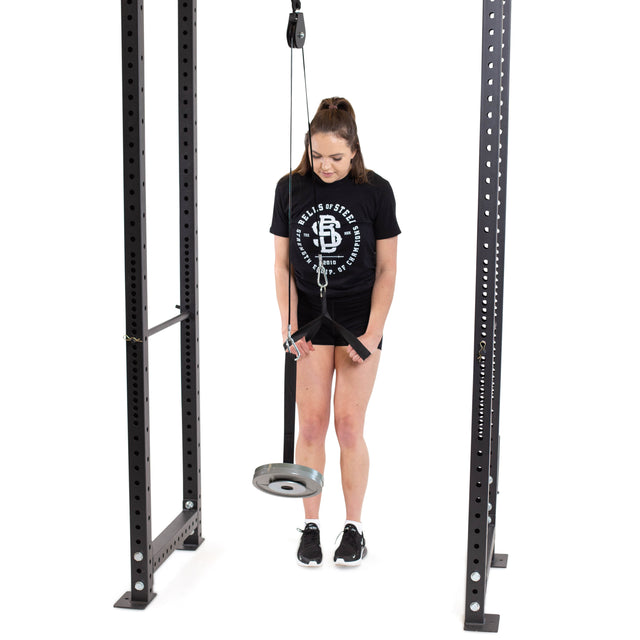 A woman in a black t-shirt and shorts skillfully performs a tricep pushdown using the Bells of Steel Cable Pulley on a weight machine. Positioned between two metal frames, she grips the bar, adjusting to its impressive weight plate capacity.