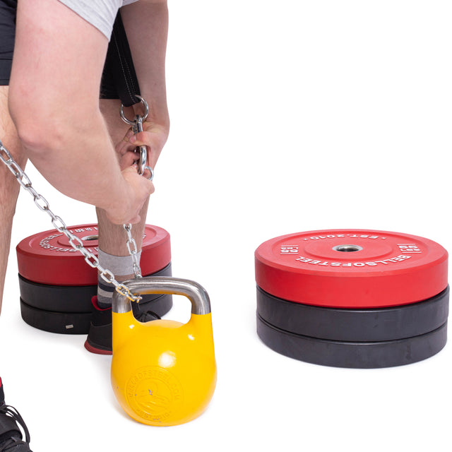 Wearing workout attire, a person attaches a chain to a yellow kettlebell using the Bells of Steel Belt Squat Belt. Red and black weight plates are stacked on the white floor, highlighting the gym's focus on weight distribution.