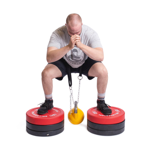 Wearing a gray shirt and black shoes, a person crouches on red weights with a kettlebell hanging from the Bells of Steel Belt Squat Belt. Their hands are clasped near their face as they master balance and focus intently on perfecting their full-body workout.