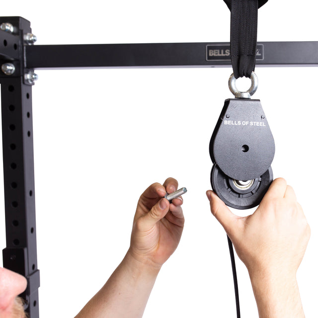 Hands are attaching a Bells of Steel Cable Pulley to a robust power rack. A small pin is being inserted for secure attachment, while a black strap links to the pulley, showcasing its impressive weight plate capacity against a crisp white background.