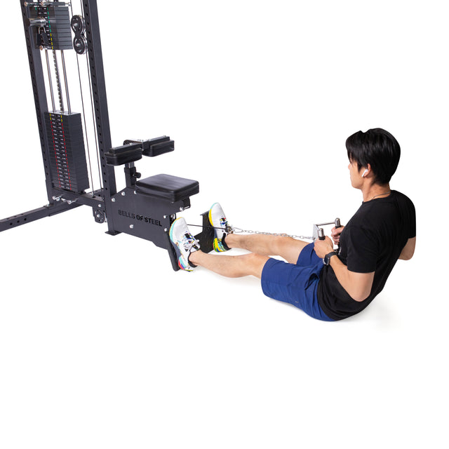 An individual exercises by performing cable rows on a low row machine. While seated, they pull a Double D Handle Cable Attachment from Bells of Steel, which is connected to the machine with a chain. The equipment harmonizes perfectly with their black shirt, blue shorts, and white sneakers.