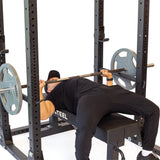 A person in black workout attire lies on a bench under a barbell with weights, doing a bench press exercise inside the Bells of Steel Hydra Flat Foot Power Rack Builder (3" x 3", ⅝" Holes) equipped with Hydra rack attachments.