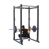 Clad in black, a person squats inside the Bells of Steel Hydra Flat Foot Power Rack Builder, a sleek and sturdy 3" x 3" black frame with modular components and numerous ⅝" holes for safely adjusting bars and hooks.