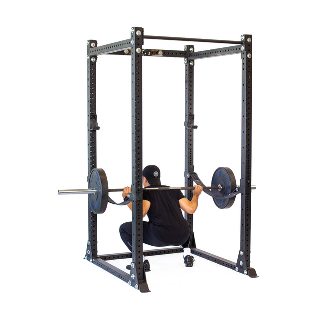 Wearing black workout clothing, a person performs a squat with a barbell on their shoulders inside the Bells of Steel Hydra Flat Foot Power Rack - Prebuilt (3" x 3", ⅝" Holes), set against a white background that highlights the gym equipment and emphasizes the exercise.