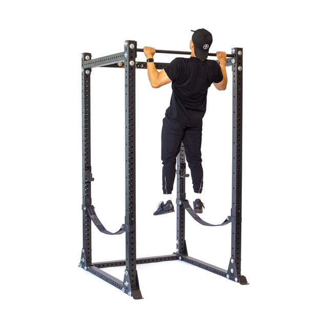 A person in black athletic wear and a cap is doing a pull-up on the Hydra Flat Foot Power Rack - Prebuilt (3" x 3", ⅝" Holes) by Bells of Steel, set against a plain white backdrop.