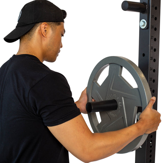 Dressed in a black t-shirt and cap, an individual adds a large Olympic-sized weight plate to the power rack. The Bells of Steel Bolt-On Plate Pegs equip the rack, which features numbered markers to indicate different height positions.