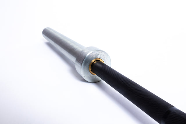 Close-up of the Bells of Steel Multi-Purpose Olympic Barbell – The Utility Bar, featuring a black and silver finish resting on a white surface. The focus is on the metal sleeve and textured grip, highlighting its quality as a premium gym equipment.