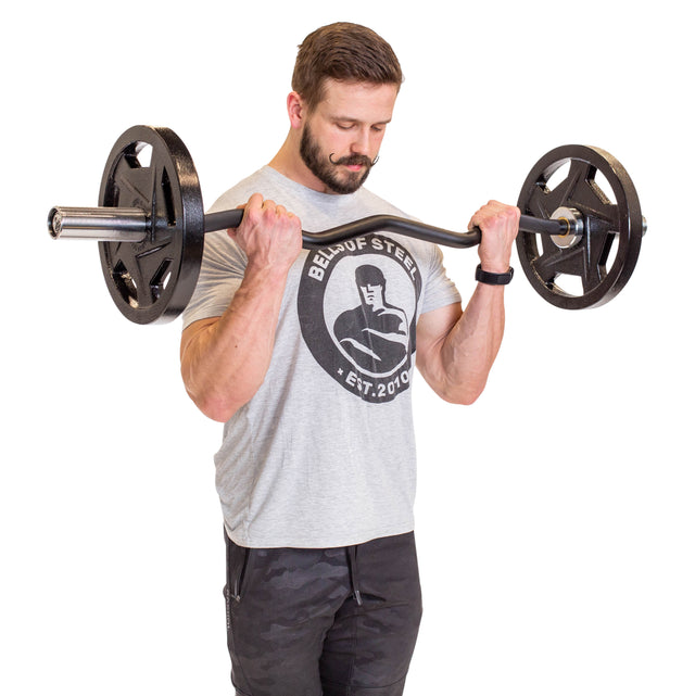 A man in a light grey shirt with a gym logo uses the Bells of Steel EZ Curl Bar (47"), loaded with weight plates. With short hair and a beard, he focuses on bicep curls to enhance his arm development against a plain white backdrop.