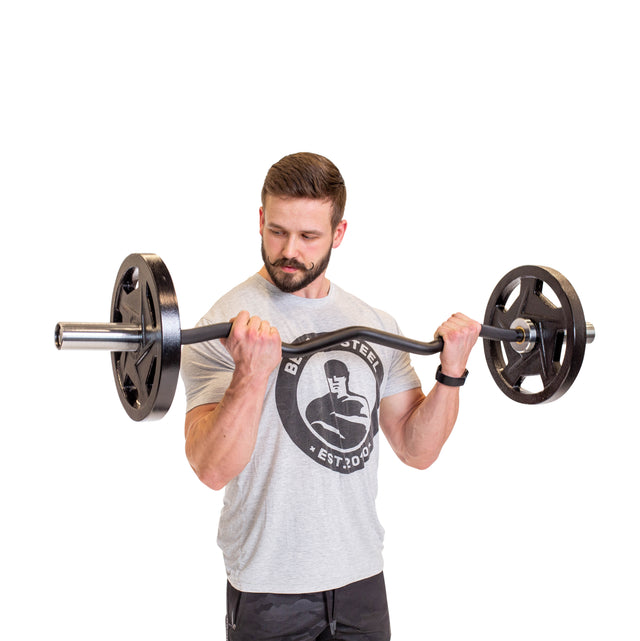 Against a plain white backdrop, a bearded man in a grey T-shirt and black shorts focuses on arm development by lifting the 47" EZ Curl Bar from Bells of Steel for bicep curls, demonstrating his dedication to strength training.