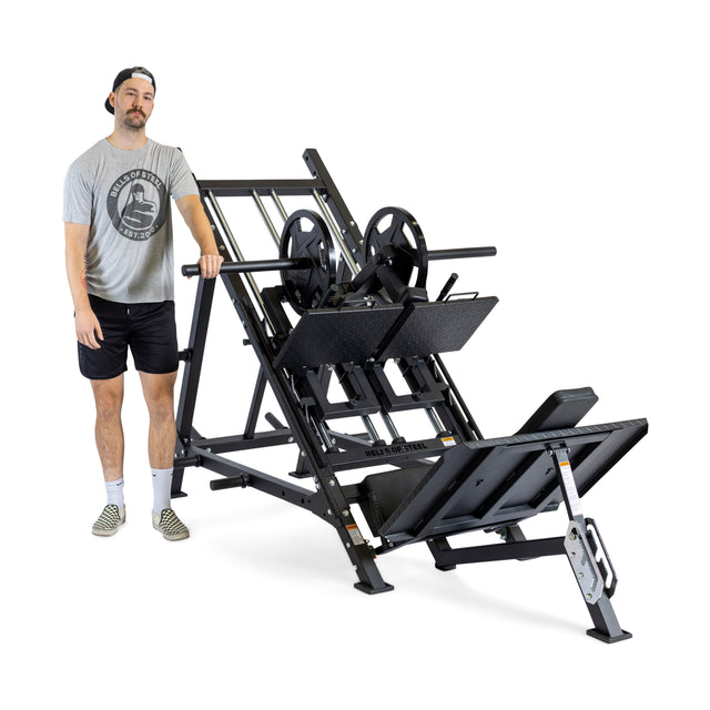 A man stands beside The Juggernaut, a black 2-in-1 Iso Leg Press & Hack Squat Machine by Bells of Steel, preparing for an intense hack squat workout. He's dressed in a gray T-shirt and black shorts, accessorized with wrist wraps and a backward cap, while sporting patterned slip-on shoes. The background is plain white.