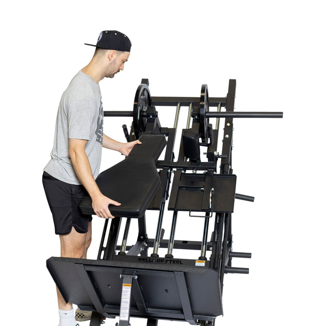 A man in a gray t-shirt and black shorts adjusts a bench on the Bells of Steel 2-in-1 Iso Leg Press & Hack Squat Machine, also known as The Juggernaut. He is wearing a black cap and standing on a white background.