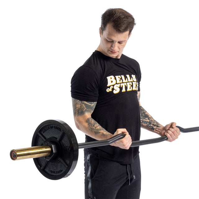 A person lifts a Rackable EZ Curl Bar for bicep curls, showcasing impressive arm development. They wear a black Bell of Steel T-shirt and black pants, with visible arm tattoos. The white background emphasizes the focus on building their biceps and triceps.