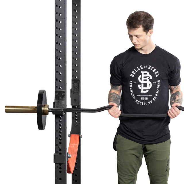 A person in a black "Bells of Steel" T-shirt and green pants holds a Rackable EZ Curl Bar with weights, standing by a squat rack, showcasing impressive arm development.