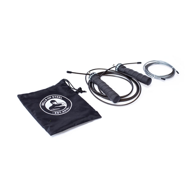 The Bells of Steel Jump Rope set, ideal for cardio, features a black rope with ergonomic gray handles, an extra silver cable, and a branded black drawstring pouch. All items are elegantly displayed on a white background.