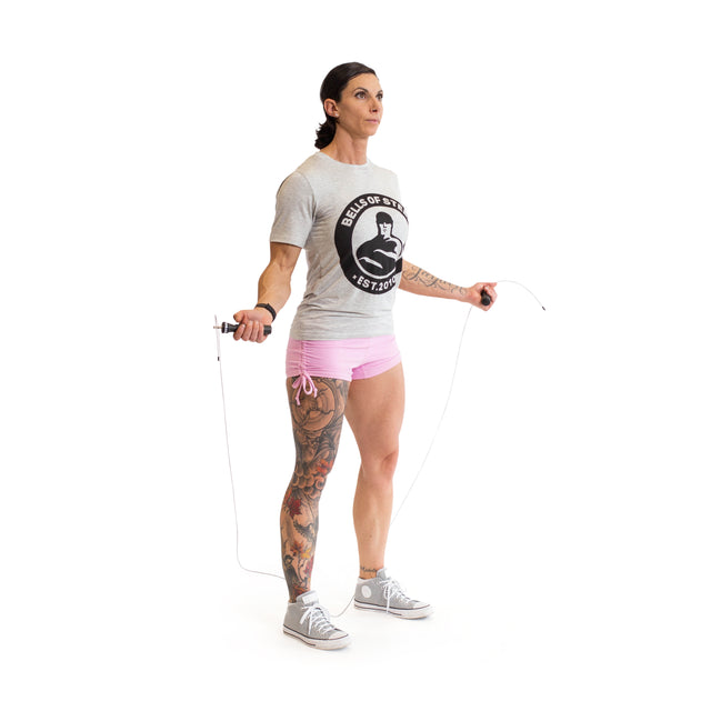 A person with a tattooed leg is doing cardio by jumping rope on a plain white background. They're wearing a gray t-shirt with a circular logo, light pink shorts, and gray sneakers, using the Bells of Steel Jump Rope that features ergonomic handles.