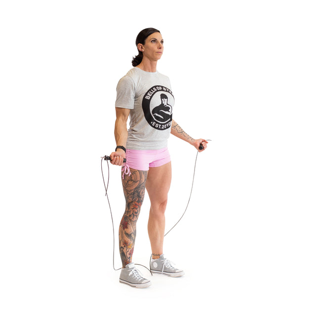 A person holds a Bells of Steel Jump Rope with ergonomic handles, clad in a gray logo t-shirt, pink shorts, and gray sneakers. A tattoo is visible on one leg against a plain white backdrop, emphasizing their readiness for cardio training.