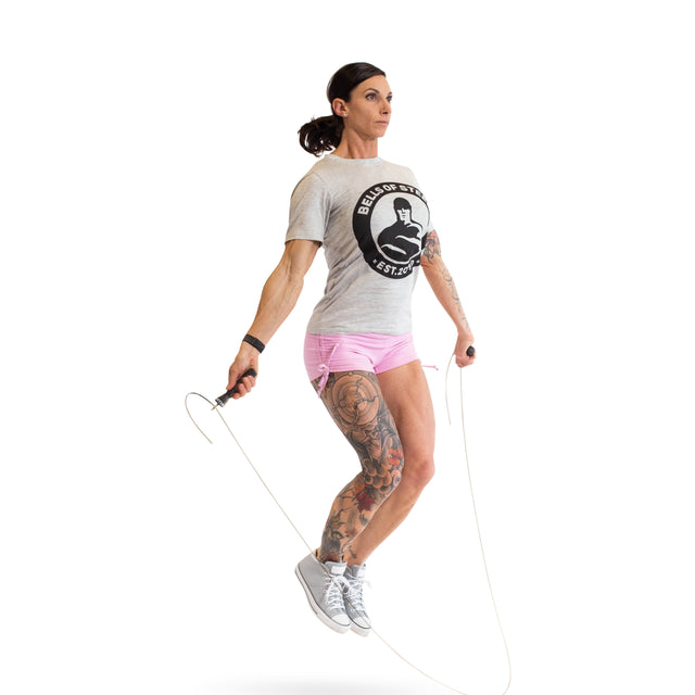 A tattooed individual in a gray T-shirt and pink shorts is mid-air while using the Bells of Steel Jump Rope for cardio training against a plain white backdrop.