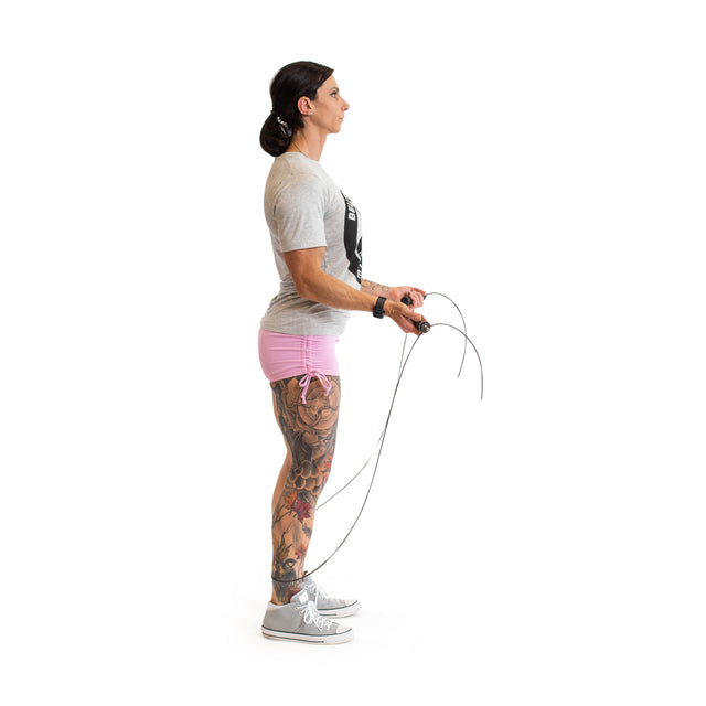 A person with long hair stands in profile, holding a Bells of Steel Jump Rope featuring ergonomic handles. They sport a gray t-shirt, pink shorts, and sneakers, ideal for cardio. Colorful tattoos decorate their right leg against a white backdrop.