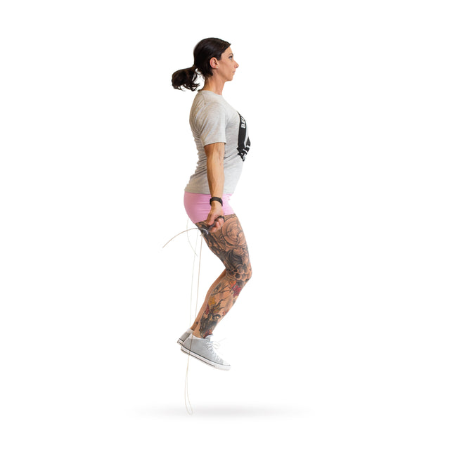 A tattooed individual jumps rope with the Bells of Steel adjustable jump rope, featuring ergonomic handles. They're dressed in a gray t-shirt, pink shorts, and sneakers against a plain white background, making this cardio training session pop.