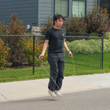 Wearing black clothes and white sneakers, a person is doing cardio by jumping rope on a sidewalk in front of a house with a black fence and green bushes, using the Bells of Steel Jump Rope.