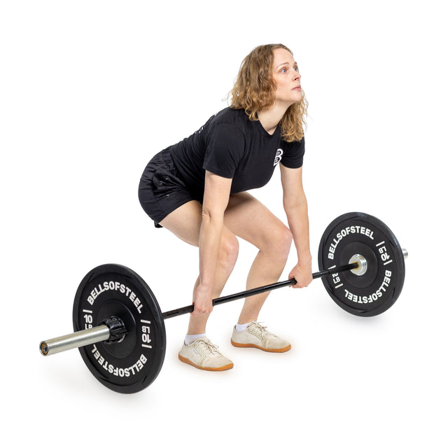 A person with smaller hands gets ready to lift a "Bells of Steel" Juno Bar - Women’s Utility Bar featuring a rust-resistant zinc coating while wearing a black shirt, shorts, and white sneakers against a pristine white background.