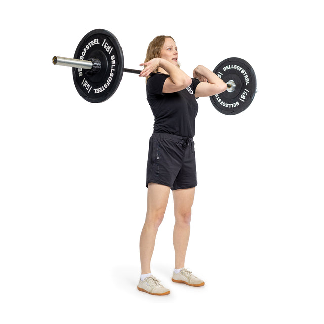 Against a plain white background, a person expertly performs a front squat using the Juno Bar - Women's Utility Bar. The bar, with rust-resistant zinc coating and "Bells of Steel" weights, is ideal for smaller hands. They wear black t-shirt and shorts with white sneakers.