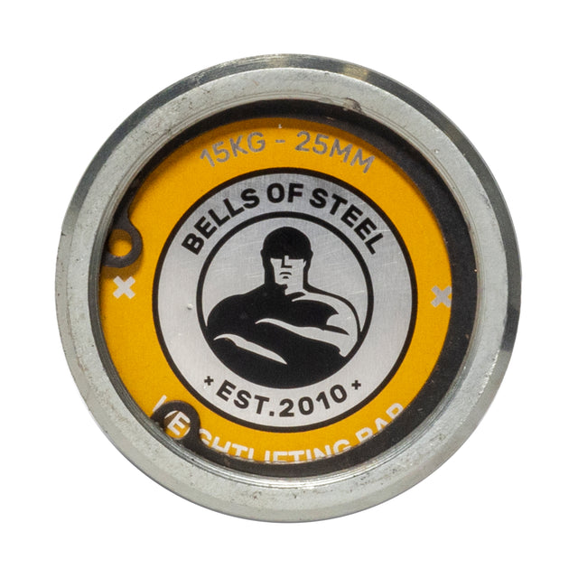 A yellow circular metal weight plate for smaller hands features the "Bells of Steel, Est. 2010" logo with a muscular figure at the center. It reads "15 KG - 25 MM." The rust-resistant zinc coating ensures durability, ideal for barbell workouts with the Juno Bar by Bells of Steel.