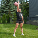 A woman stands on grass holding a pink kettlebell from the Bells of Steel Ultimate Backyard Training Bundle overhead with one arm, dressed in a black shirt, shorts, and white sneakers. Trees, a fence, and a building in the background create the perfect setting for an outdoor workout.