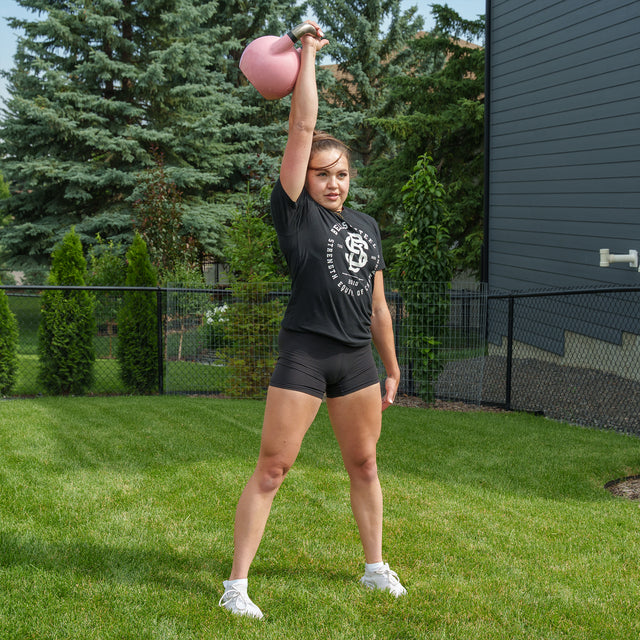 A woman stands on grass holding a pink kettlebell from the Bells of Steel Ultimate Backyard Training Bundle overhead with one arm, dressed in a black shirt, shorts, and white sneakers. Trees, a fence, and a building in the background create the perfect setting for an outdoor workout.