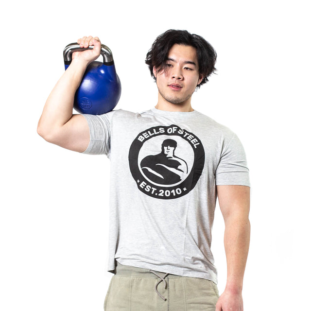 A person with medium-length dark hair is holding a blue Competition Kettlebell by Bells of Steel on their shoulder. They are dressed in a gray T-shirt featuring the "Bells of Steel Est. 2010" logo and light-colored pants, standing against a white background to highlight the kettlebell's ergonomic design, ideal for sport enthusiasts.