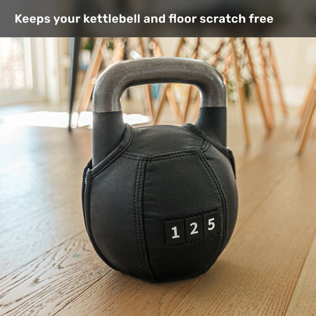 A black kettlebell featuring the Bells of Steel Kettlebell Wrap, stitched and marked “125,” rests on a wooden floor. Text above states, “Keeps your kettlebell and floor scratch free for optimal floor protection.” Sunlight pours in through windows behind.