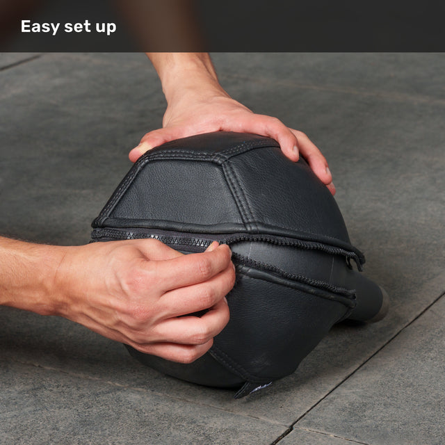 A person zips up a black, multi-paneled Bells of Steel Kettlebell Wrap on a gray floor, providing extra floor protection. The text “Easy set up” appears at the top left on a dark overlay.