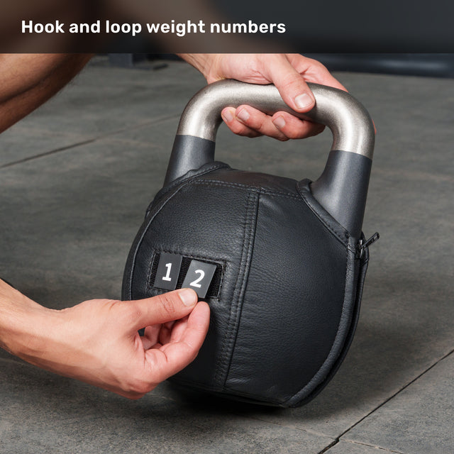 A “12” weight label is secured to a Bells of Steel Kettlebell Wrap—a black, adjustable competition kettlebell accessory with a hook-and-loop patch—featuring a metallic handle and resting on a concrete floor. Designed for versatility and floor protection, the text above reads: “Hook and loop weight numbers.”.