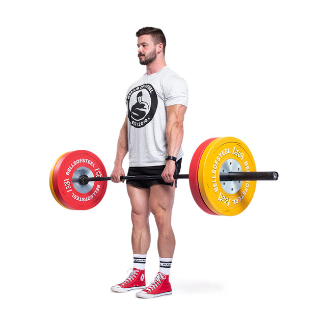 A man in a white t-shirt and black shorts performs a deadlift with a barbell, loaded with Bells of Steel KG Competition Bumper Plates in bold red and yellow. He is wearing red high-top sneakers, standing on a white floor against a crisp white background.