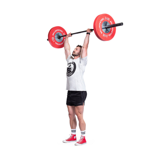 A man in a white shirt and black shorts is performing an overhead lift with a barbell equipped with Bells of Steel KG Competition Bumper Plates. His red sneakers complement the vibrant weights as he stands against a white background, looking focused and engaged in his Olympic lifting routine.